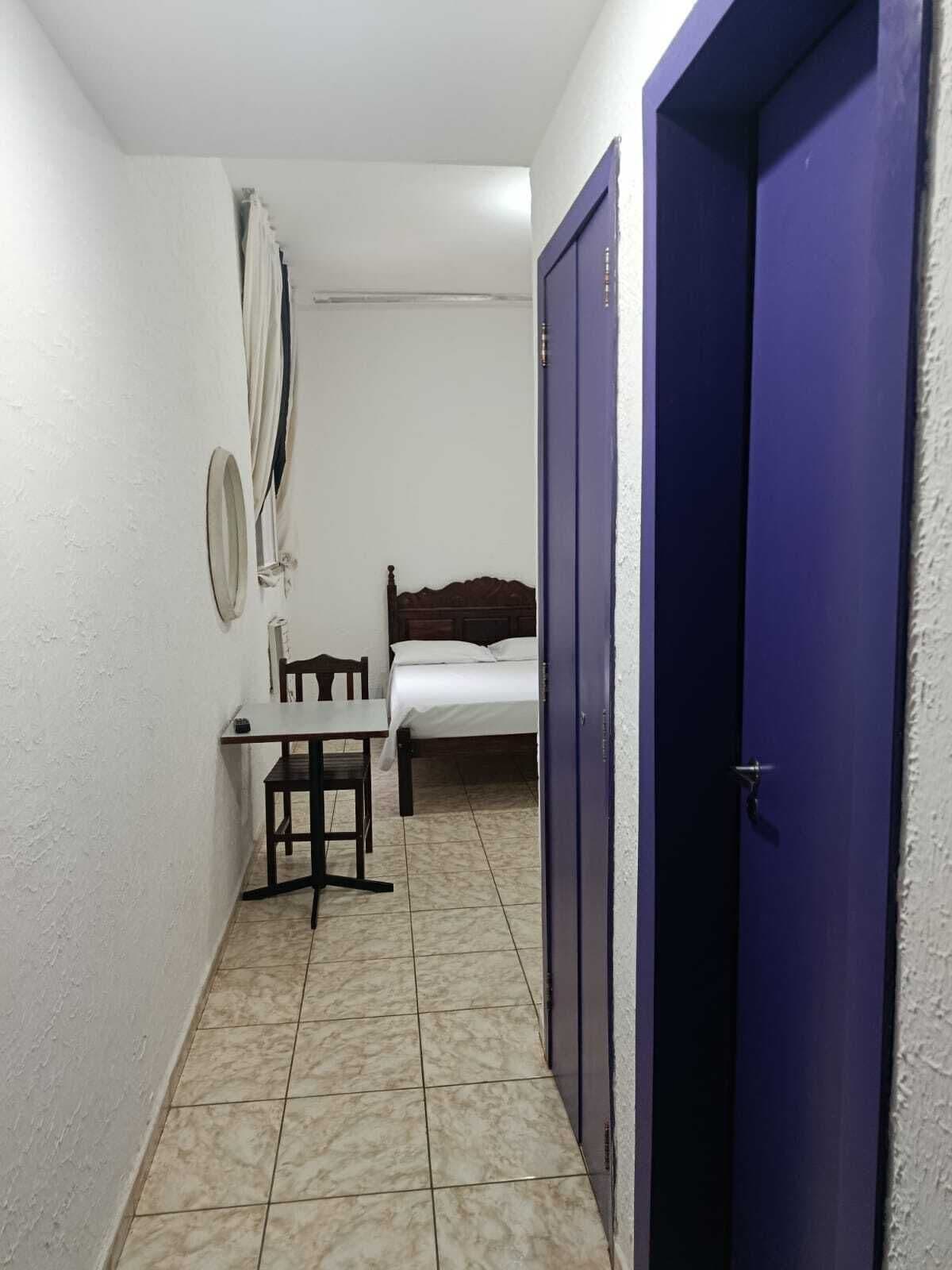 A narrow hallway with a bed, a small table with a chair, and a blue door.