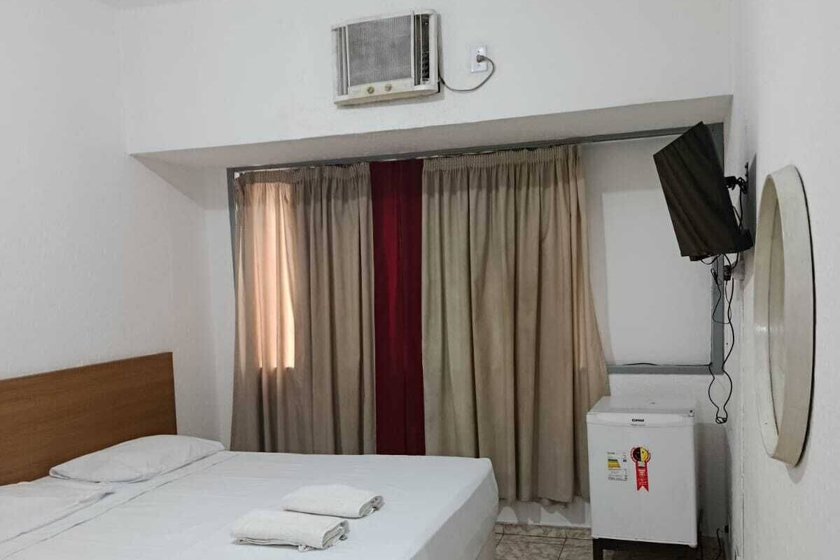 A hotel room with a bed, a small refrigerator, an air conditioning unit, and a television mounted on the wall.