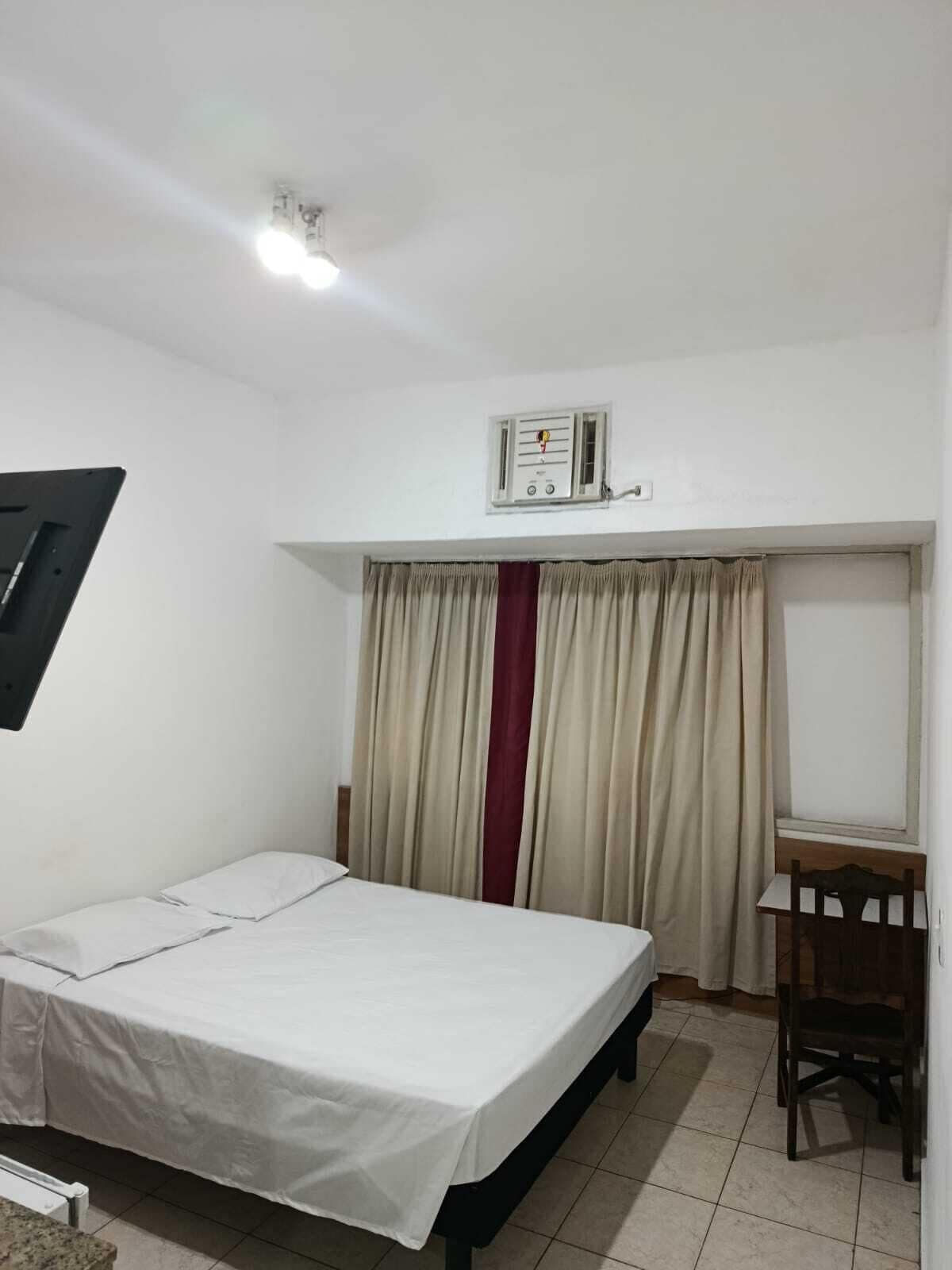 Quarto pequeno e limpo, com uma cama de solteiro, uma cadeira, uma mesa e uma janela com cortinas.