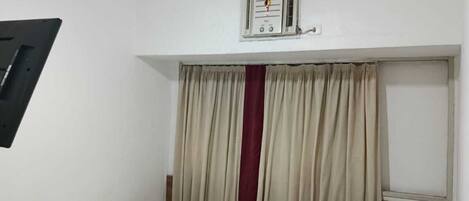 A small, clean room with a single bed, a chair, a desk, and a window with curtains.
