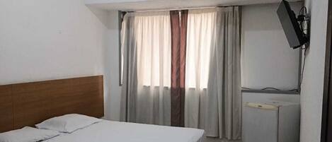 Quarto de hotel com cama, escrivaninha, televisão e janela com cortinas.