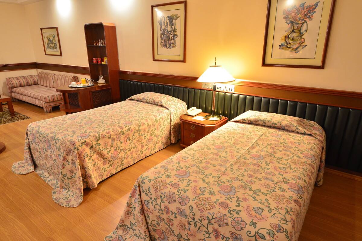 Ein Hotelzimmer mit zwei Einzelbetten, einem Sofa, einem kleinen Tisch mit Lampe und einem Bücherregal.