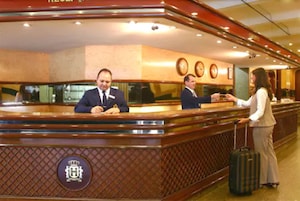 Una reception con due membri dello staff, una donna con una valigia e un orologio a parete.