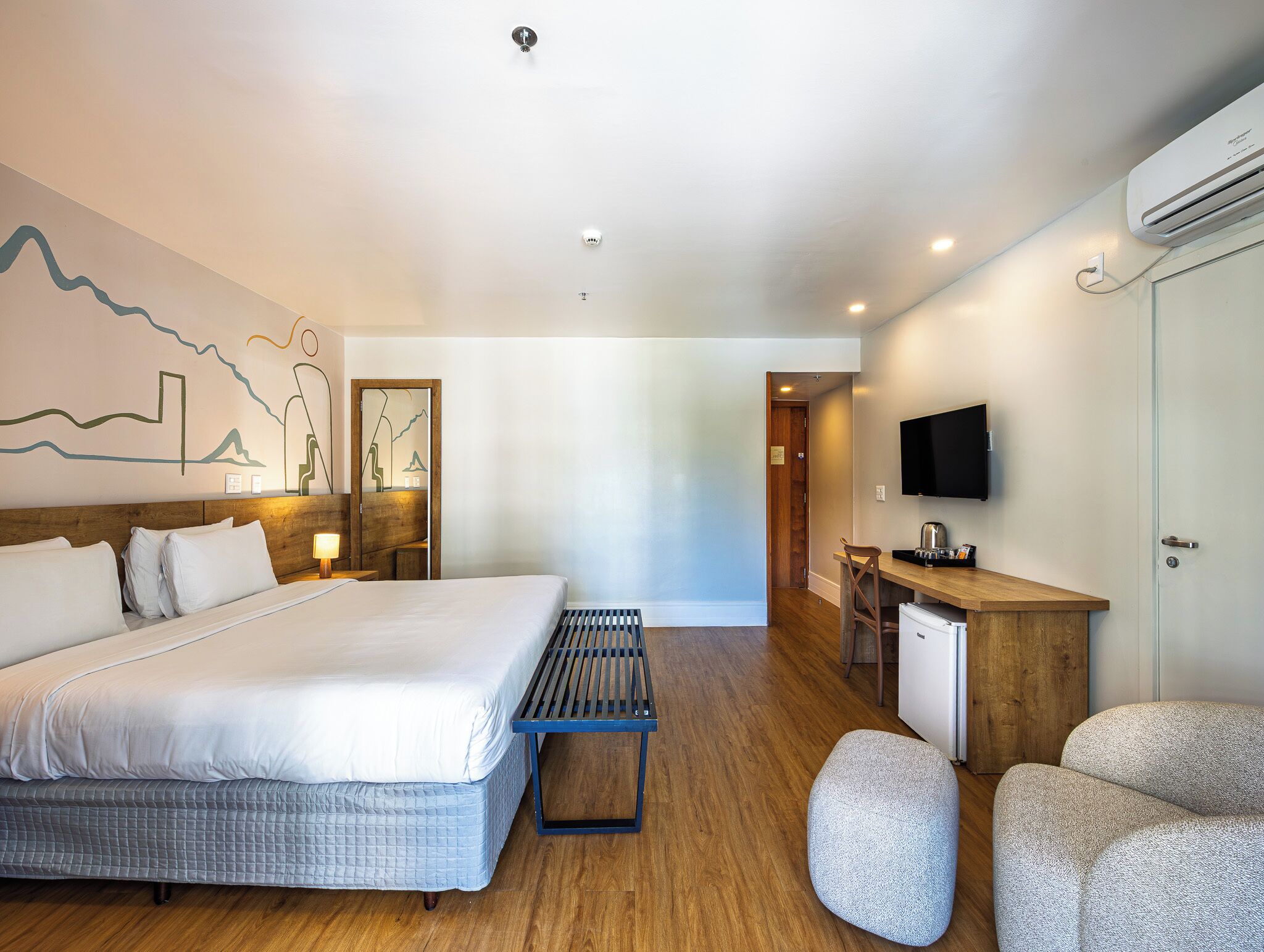 A hotel room with a bed, a desk, a chair, a TV, and an air conditioning unit.
