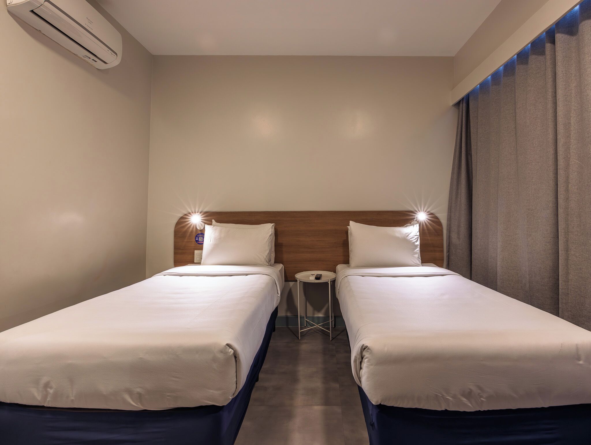 Habitación de hotel con dos camas individuales, una mesita redonda, un equipo de aire acondicionado y una cortina.
