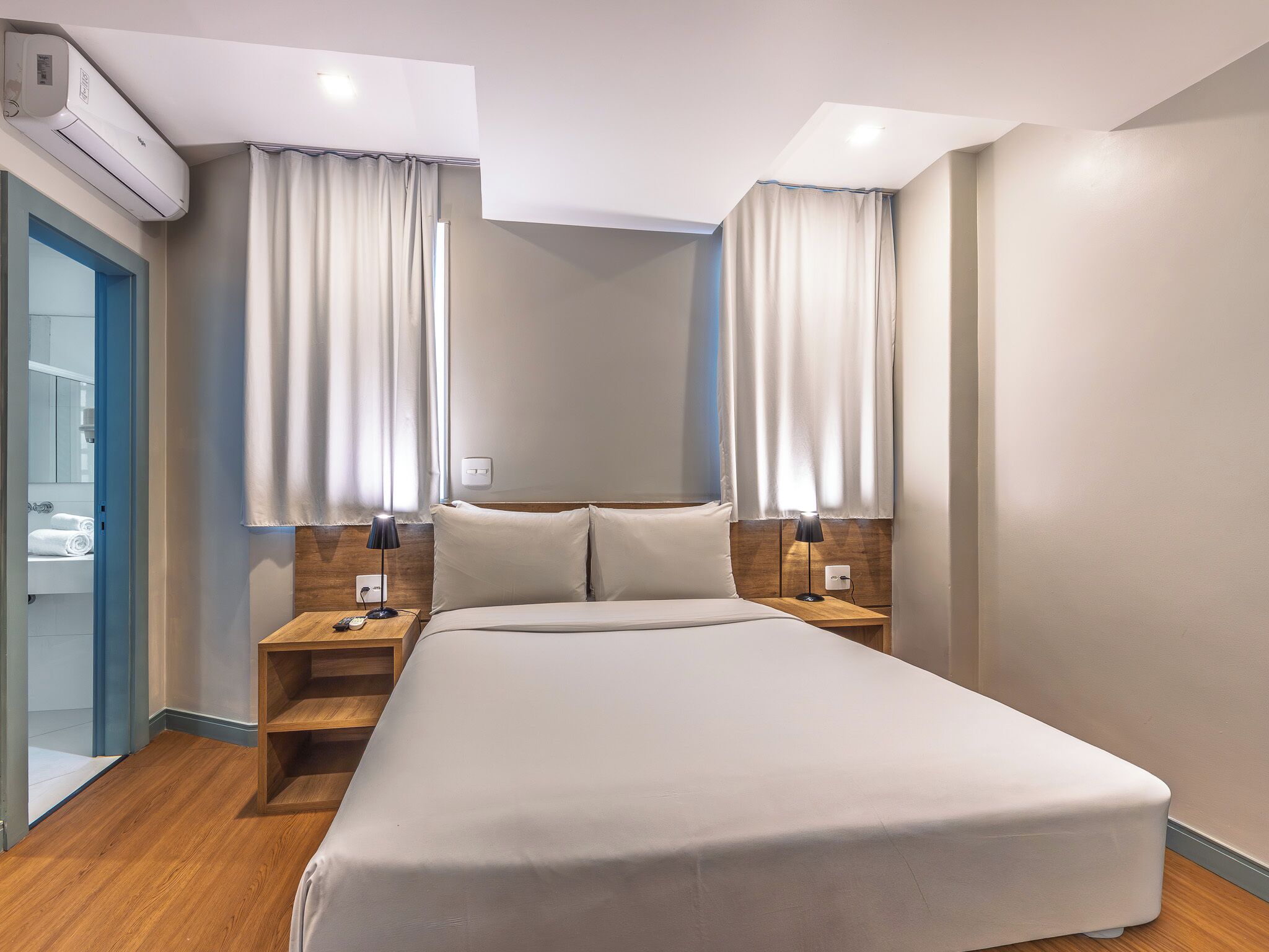 A hotel room with a bed, two bedside tables, a nightstand, an air conditioning unit, and a bathroom visible through an open door.