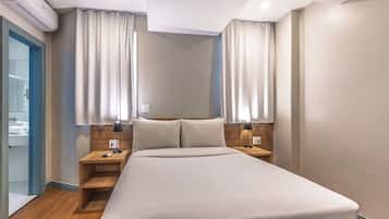 A hotel room with a bed, two bedside tables, a nightstand, an air conditioning unit, and a bathroom visible through an open door.