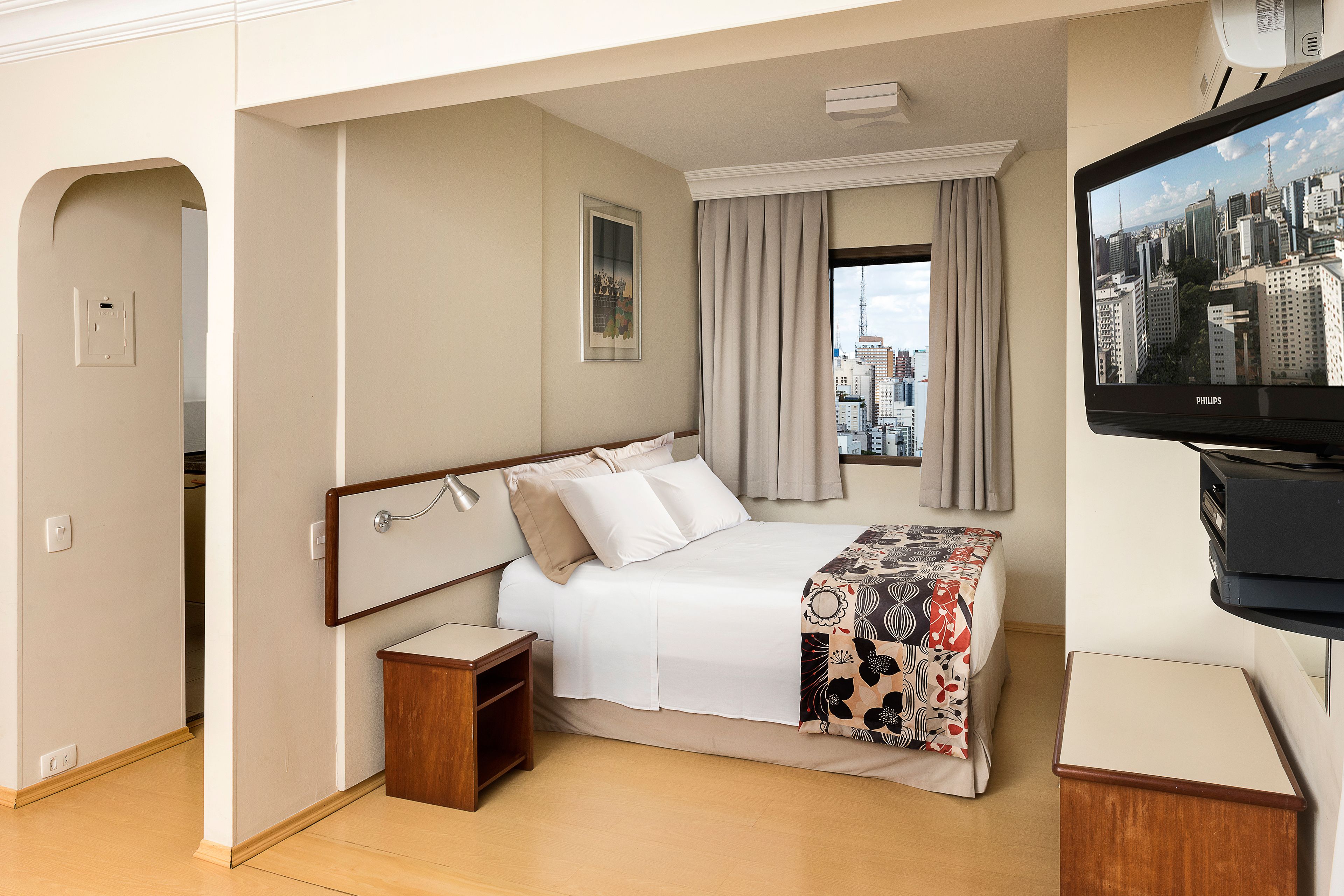A hotel room with a bed, a small bedside table, a TV, and a window with a cityscape view.