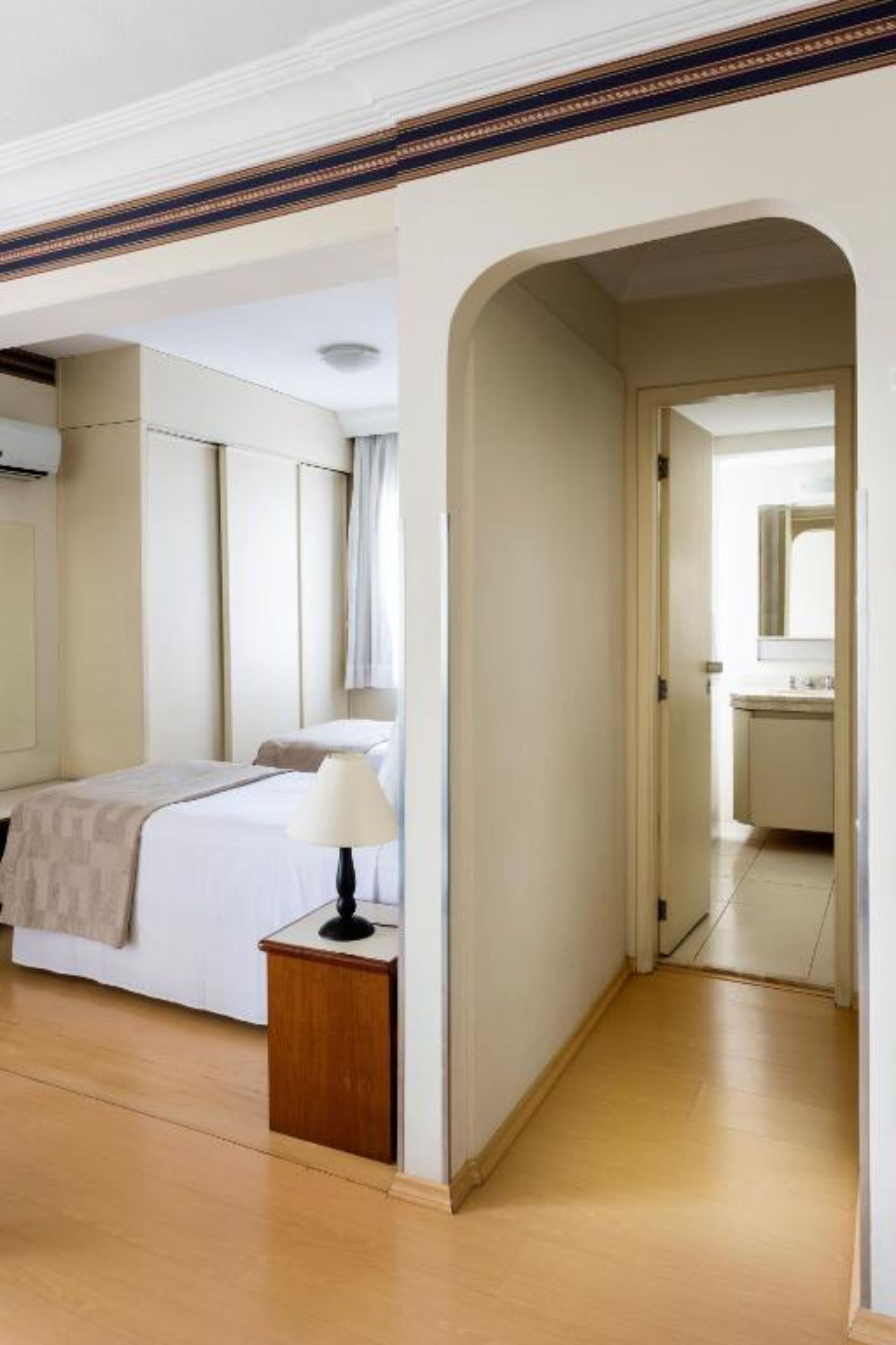 Quarto de hotel com cama, mesa de cabeceira, abajur e vista para o banheiro através de um arco.