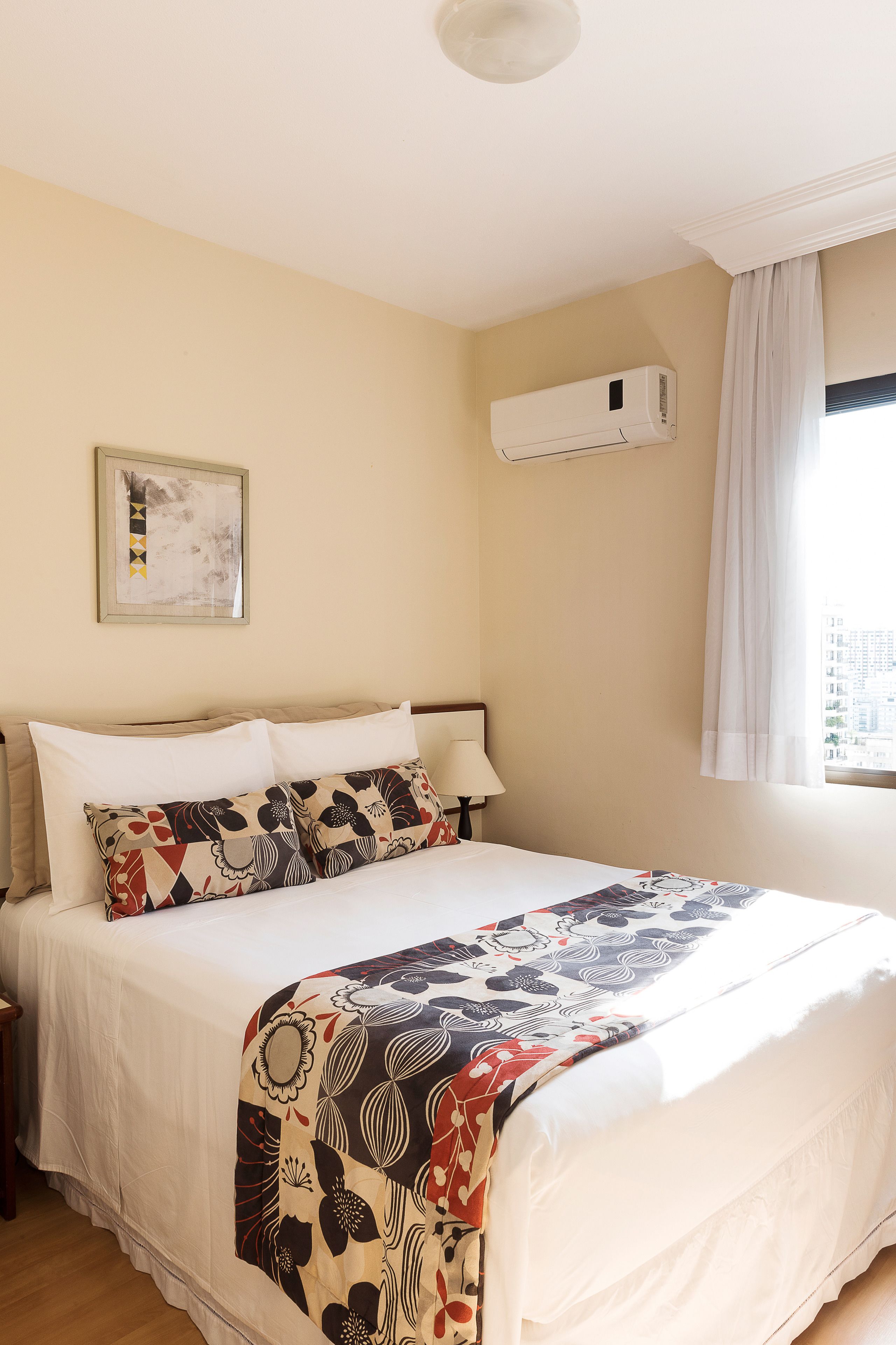 A hotel room with a bed, a nightstand, a lamp, an air conditioning unit, a window with curtains, and a framed picture on the wall.
