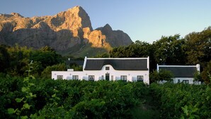 A white building with a black roof, surrounded by lush greenery and a mountain in the background. - Klein Waterval Riverside Lodge (Franschhoek)