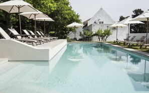 A swimming pool with lounge chairs and umbrellas, surrounded by trees and a building with a white facade.