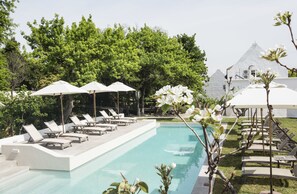 Area piscina con lettini e ombrelloni, circondata da alberi e un edificio bianco sullo sfondo.