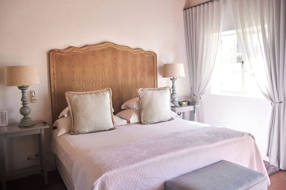 A neatly made bed with two pillows, a bedside table with a lamp, and a window with curtains.