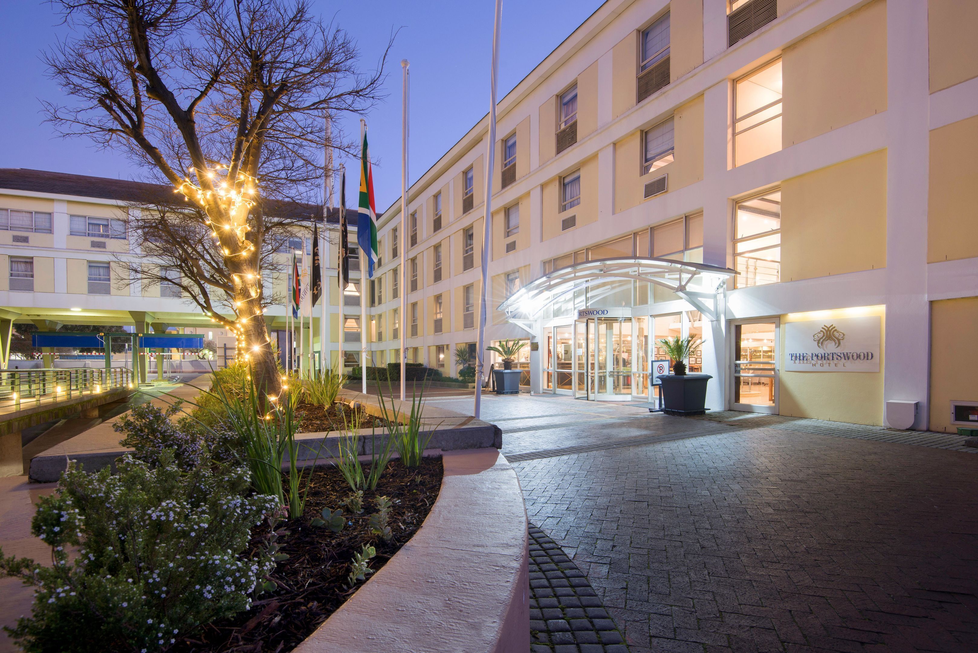 A hotel entrance with a glass canopy, a sign for The Portswood Hotel, and a landscaped area with plants and a tree.