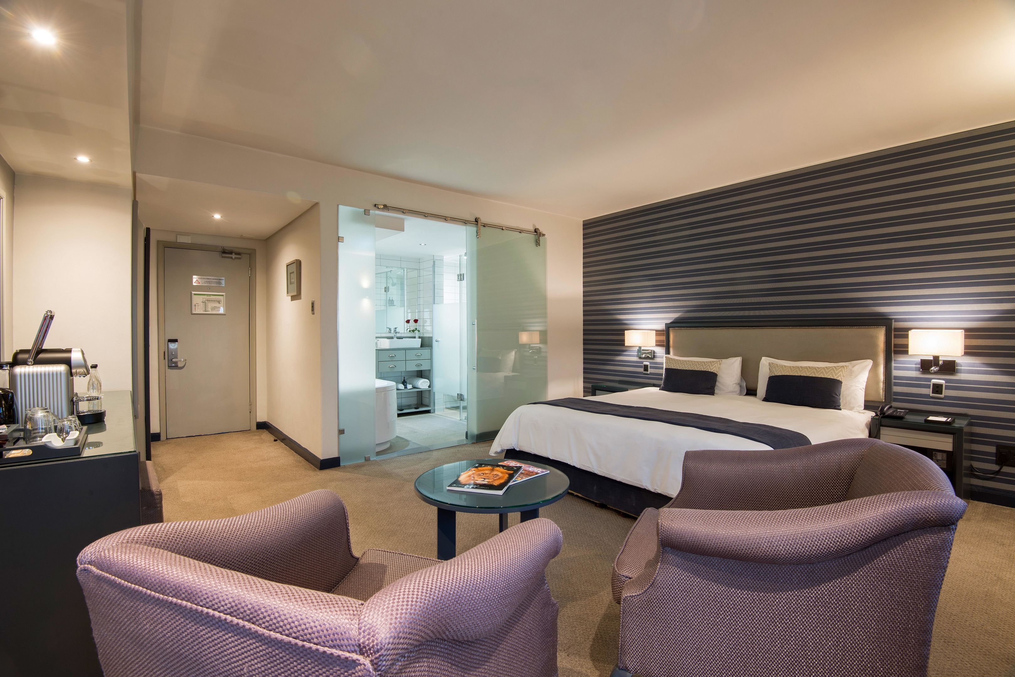 A modern hotel room with a large bed, two armchairs, a small table with a magazine, and a bathroom visible through a glass door.