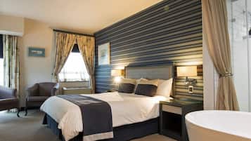 A modern hotel room with a large bed, a sitting area with a chair, a bedside table with a lamp, and a large bathtub.