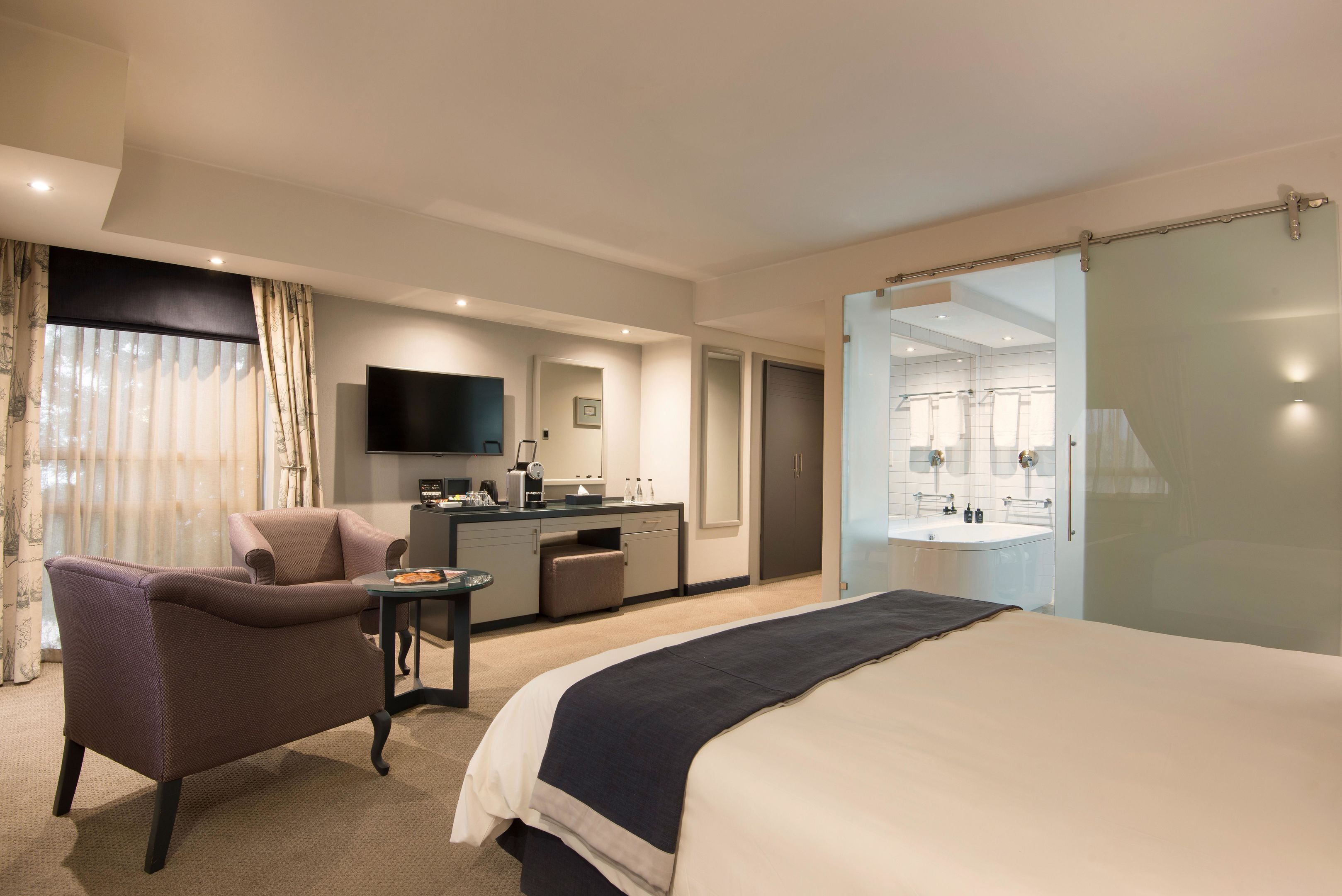 Quarto de hotel com cama, escrivaninha, cadeira, televisão, banheiro com pia e espelho, e porta de correr.
