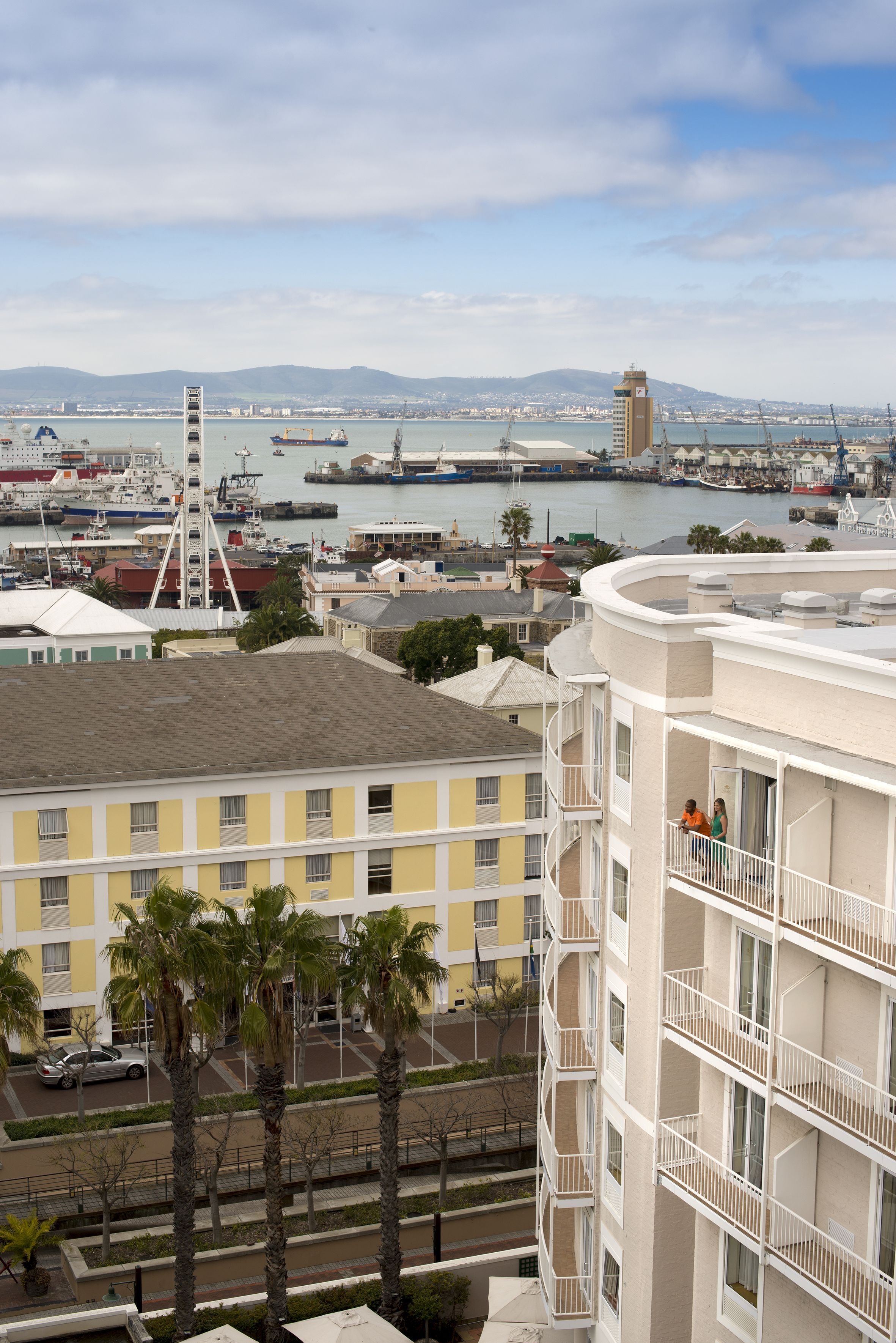 A high-rise building with balconies overlooking a harbor with boats and a distant mountain range.