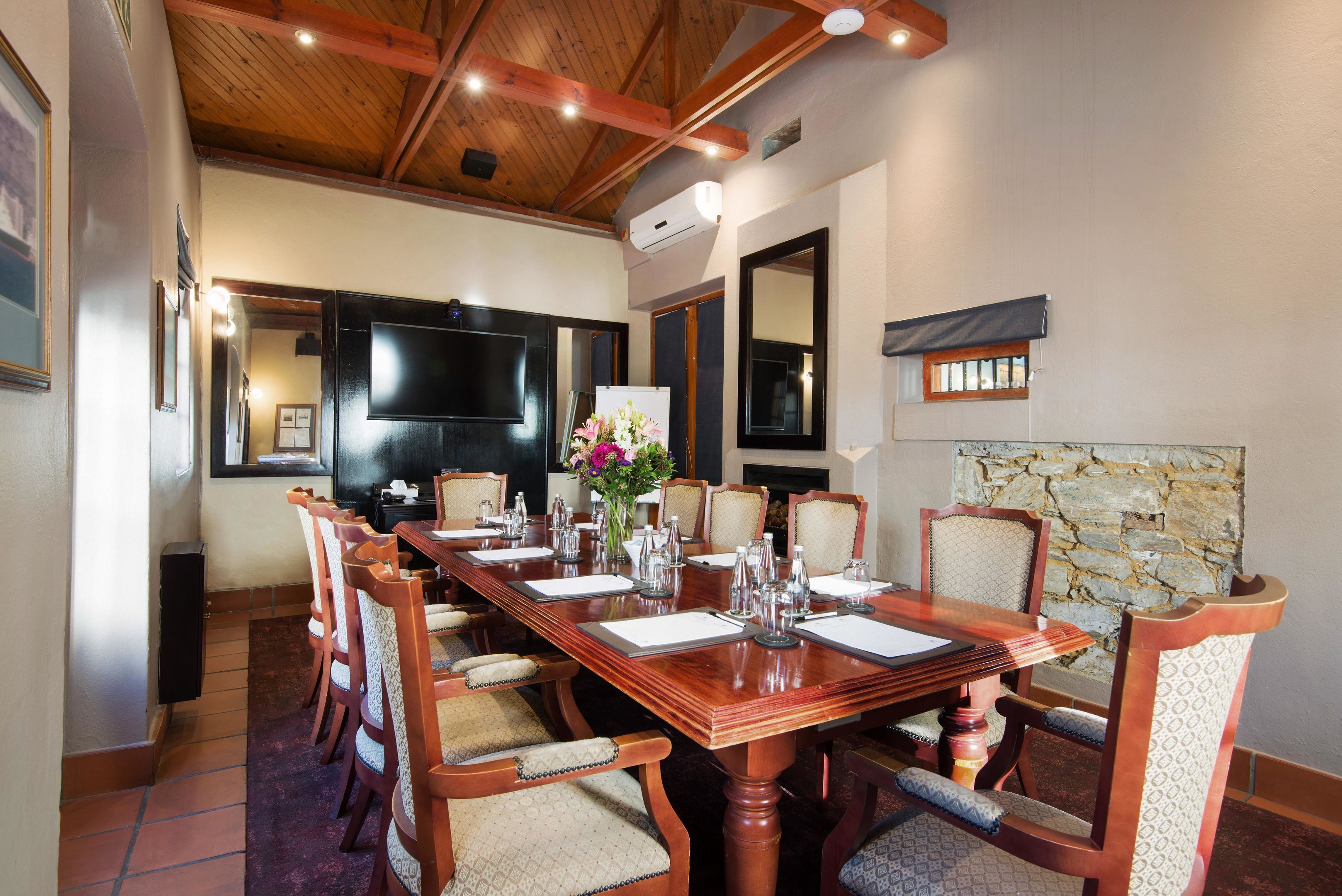 A conference room with a long wooden table, multiple chairs, a large flat-screen TV, and a stone wall.