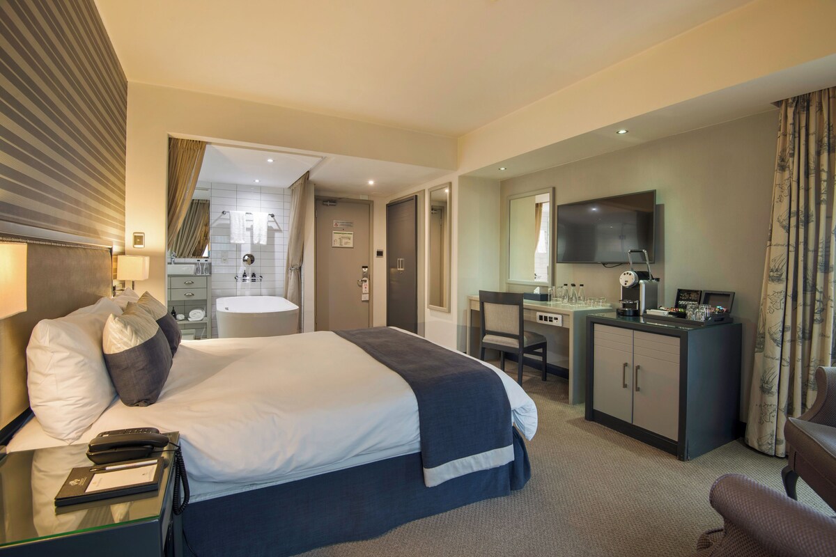 Habitación de hotel con una cama grande, un escritorio con computadora, un espejo y un baño visible a través de una puerta abierta.