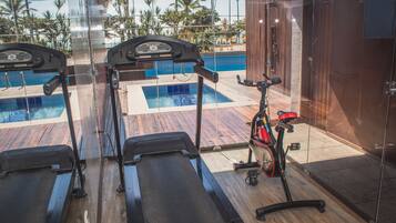 Un gimnasio con dos cintas de correr y una bicicleta estática, con vistas a una piscina y palmeras al fondo.