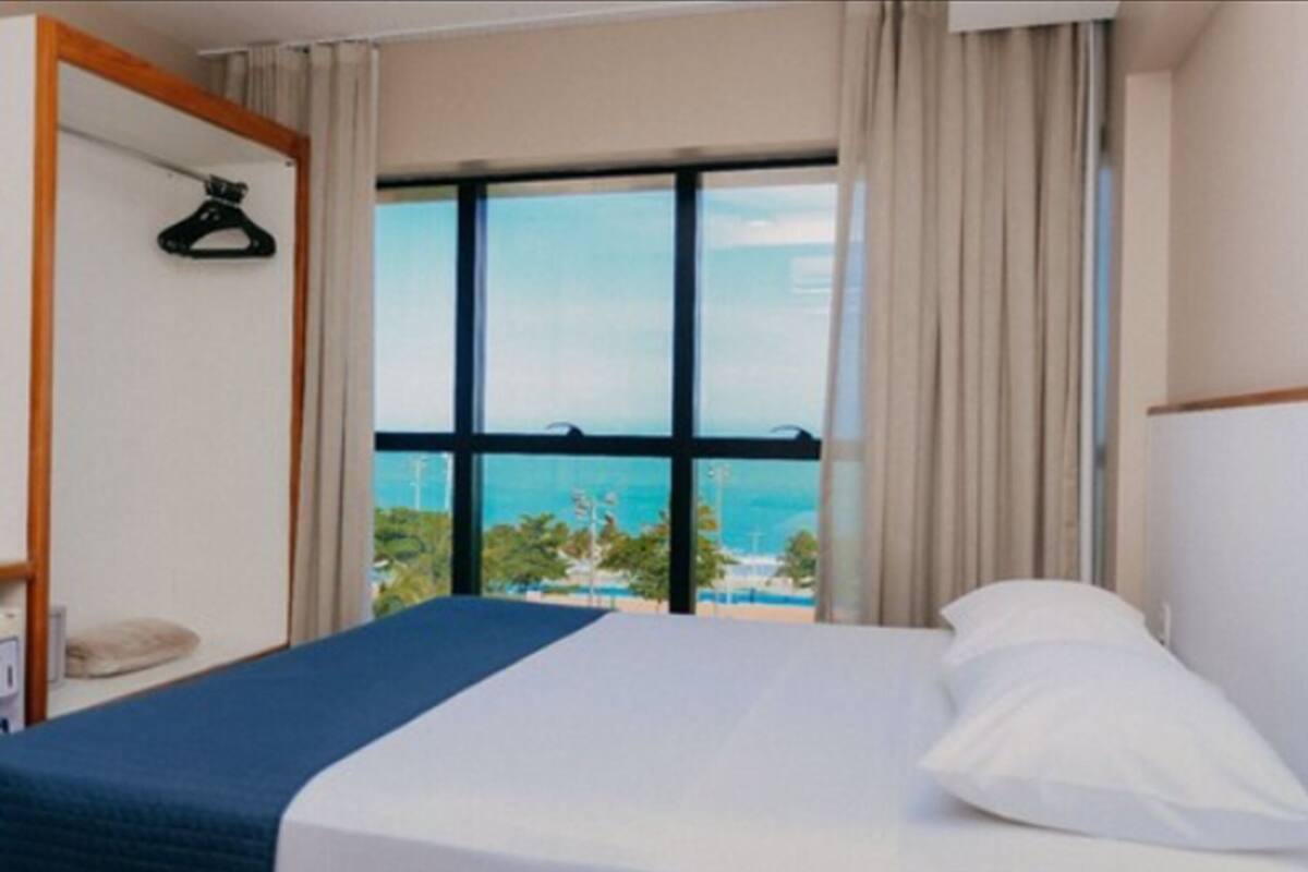 A hotel room with a large window, a bed, a hanging coat rack, and a view of the sea.