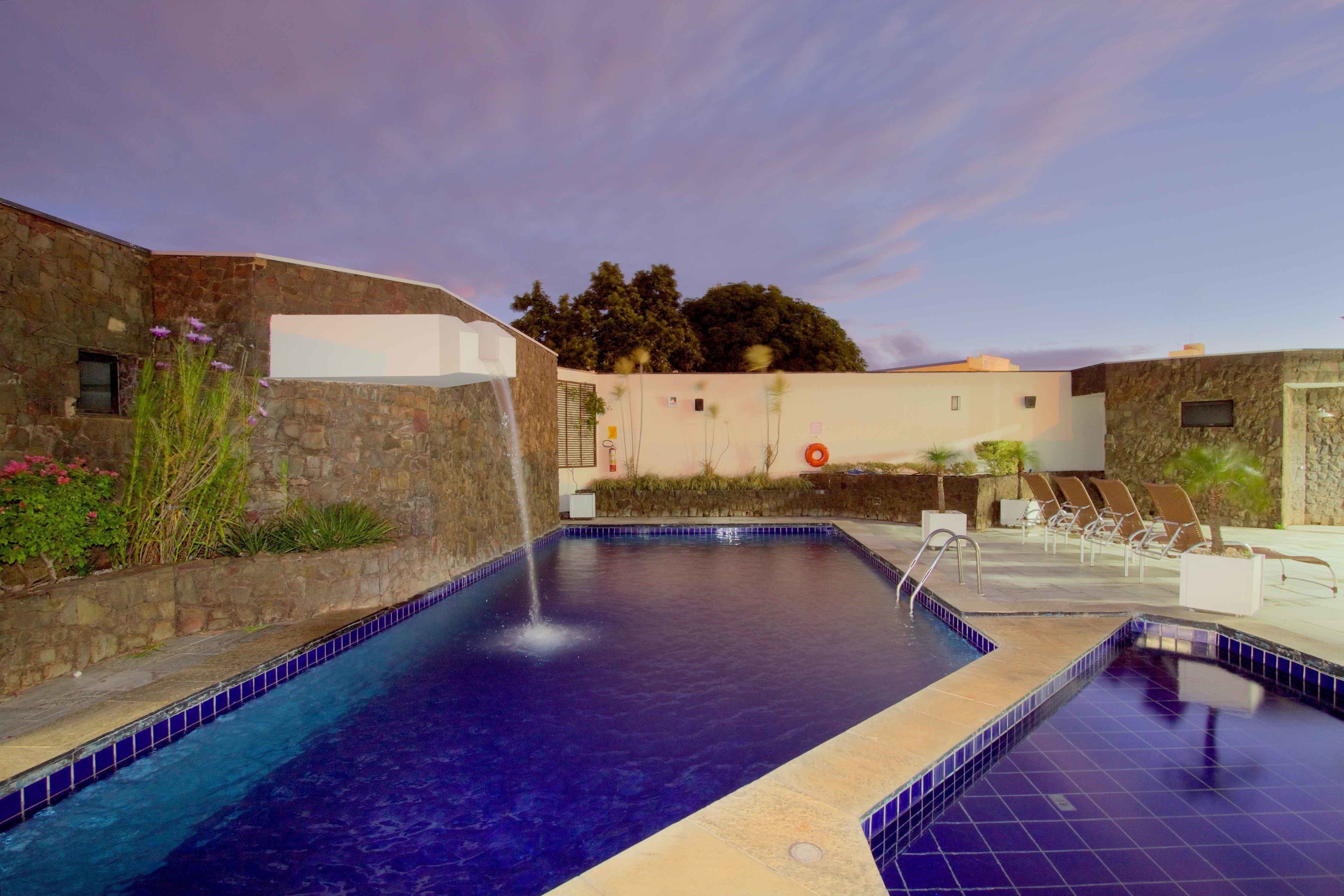 Una piscina con caída de agua, rodeada de sillas de descanso y un muro de piedra.