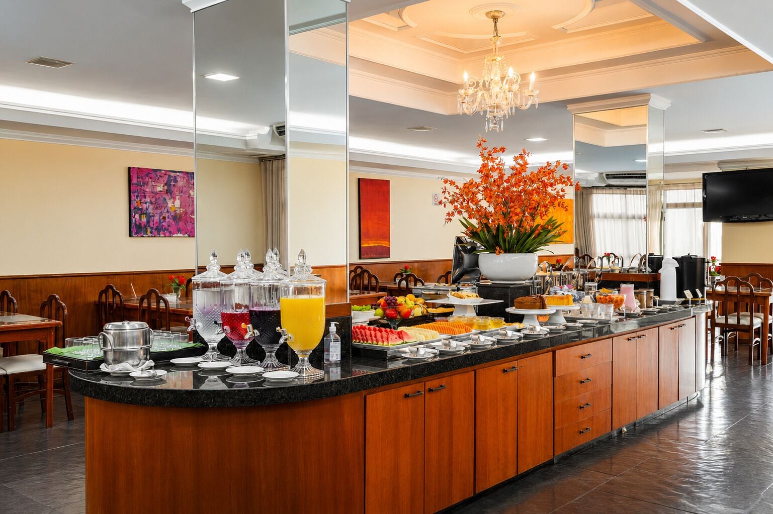 A breakfast bar with various food items and beverages, a chandelier, and a television in a hotel setting.