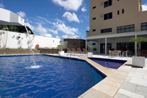Piscina com atração de água, área de estar ao ar livre e um edifício de hotel ao fundo.