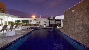 Una piscina con sillas de descanso y un muro de piedra, rodeada de edificios con un cartel que dice 'Academia'.