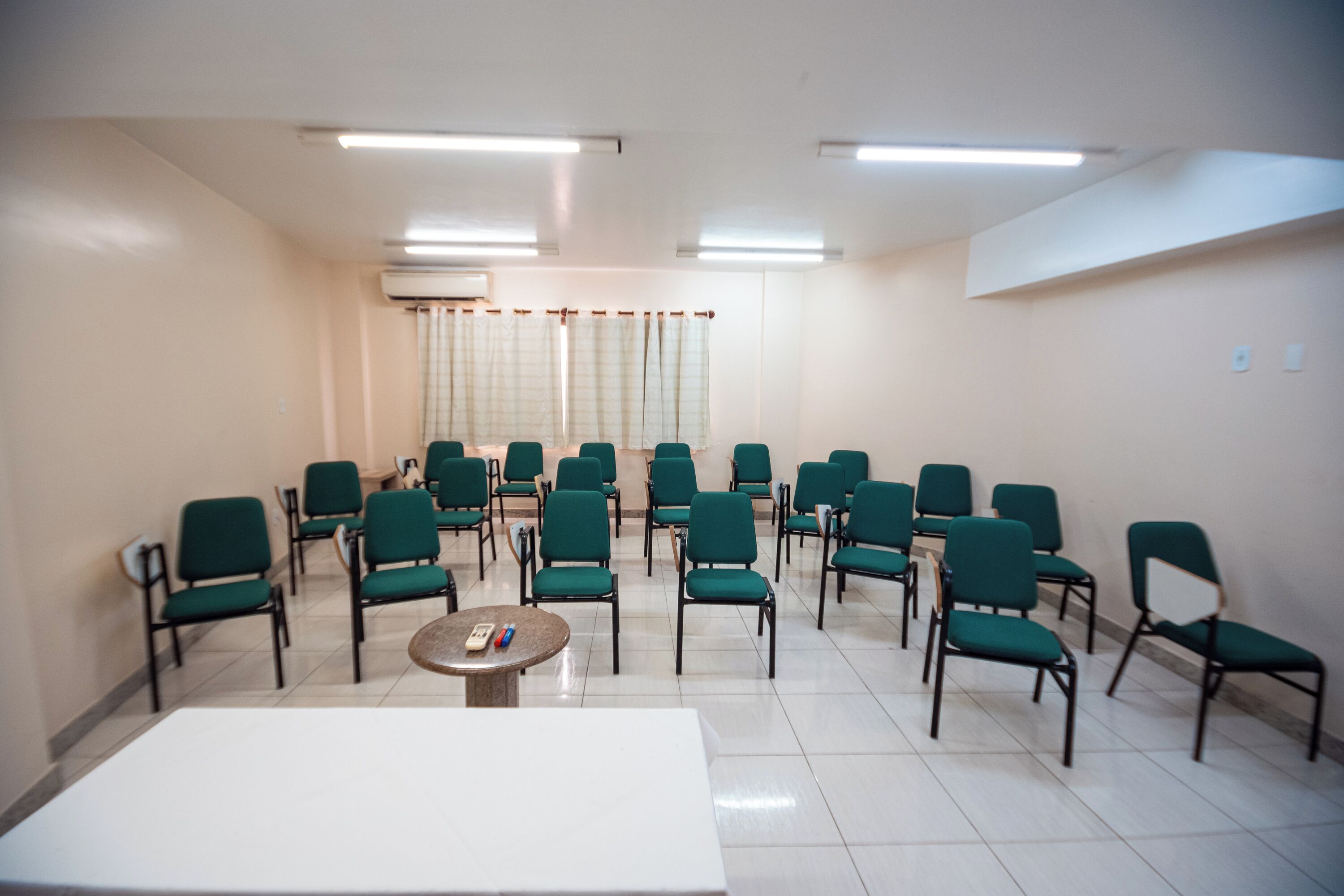 Un salón de conferencias con filas de sillas verdes, una mesa blanca e iluminación fluorescente en el techo.