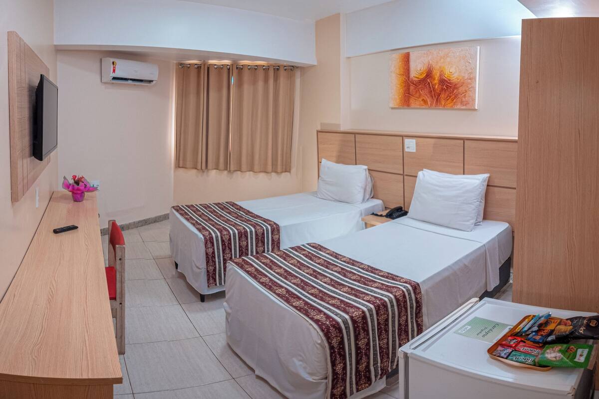 A hotel room with two beds, a desk, a TV, an air conditioner, and a painting on the wall.