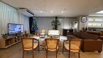Un salon avec un téléviseur, un sapin de Noël, des fauteuils en osier, un canapé, une horloge et une plante.