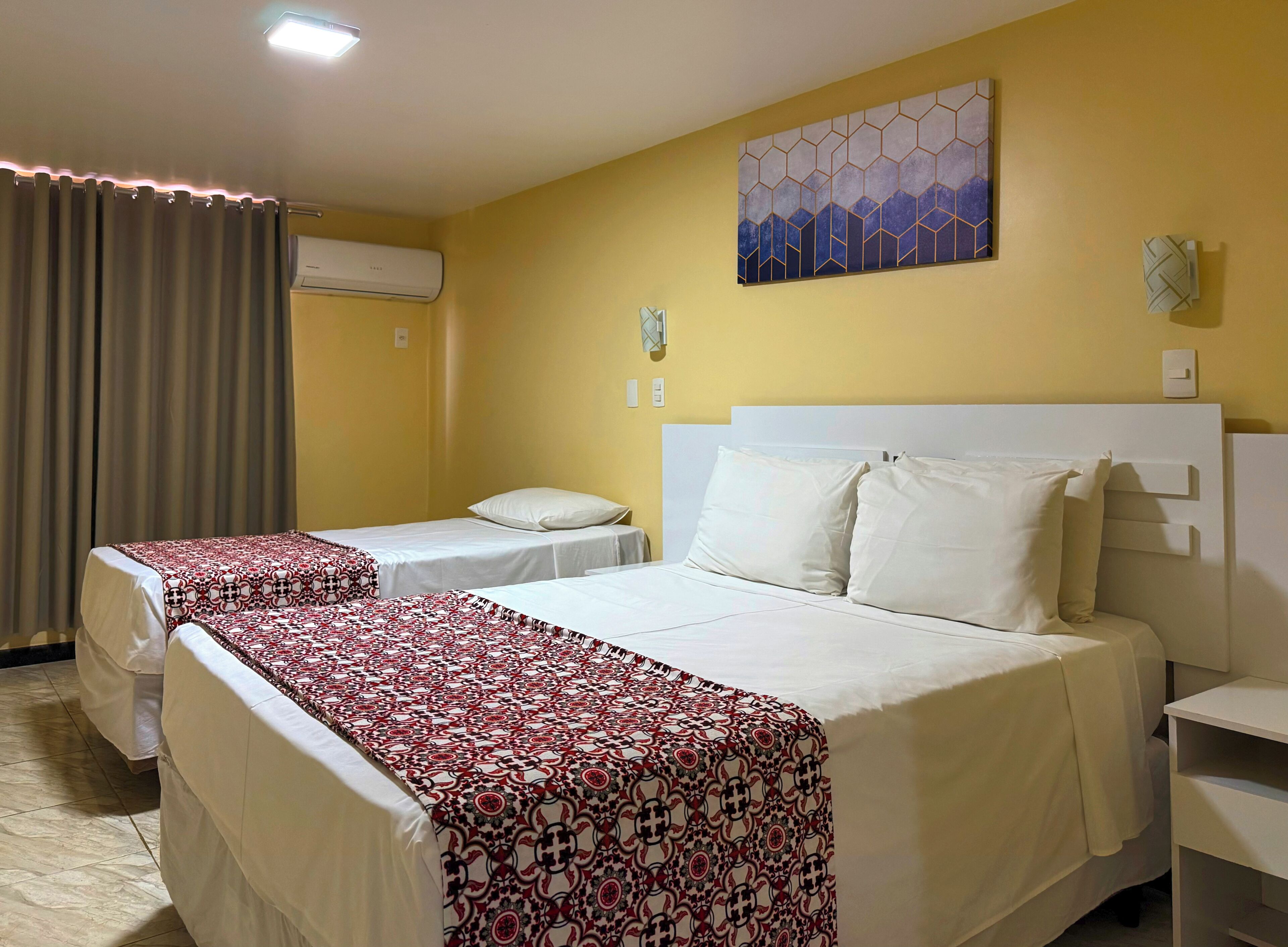 A hotel room with two beds, a large window with curtains, a wall-mounted air conditioner, and a geometric painting.