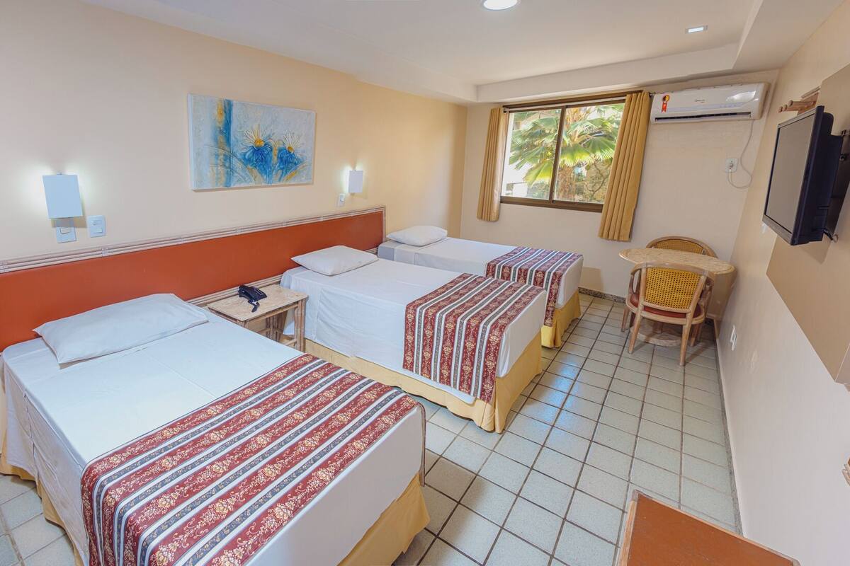 A hotel room with two single beds, a small table, a chair, a TV, and a window with curtains.