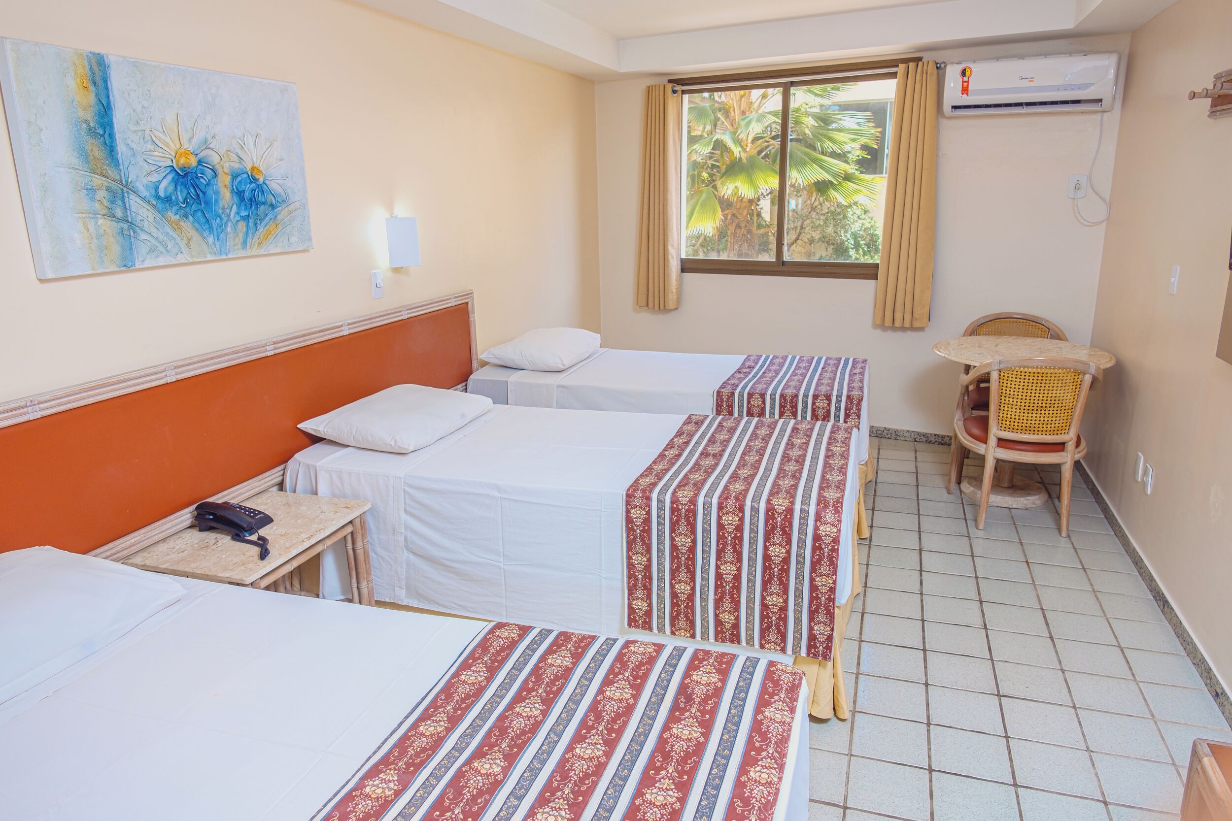 Une chambre d’hôtel avec deux lits simples, une petite table avec une chaise, un tableau au mur et un appareil de climatisation.