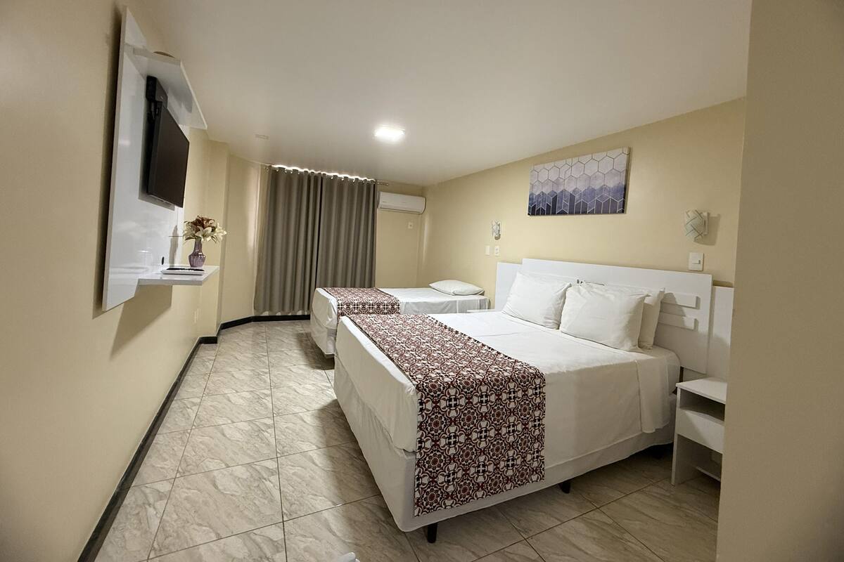 A hotel room with a bed, a bedside table, a TV mounted on the wall, a window with curtains, and a painting on the wall.