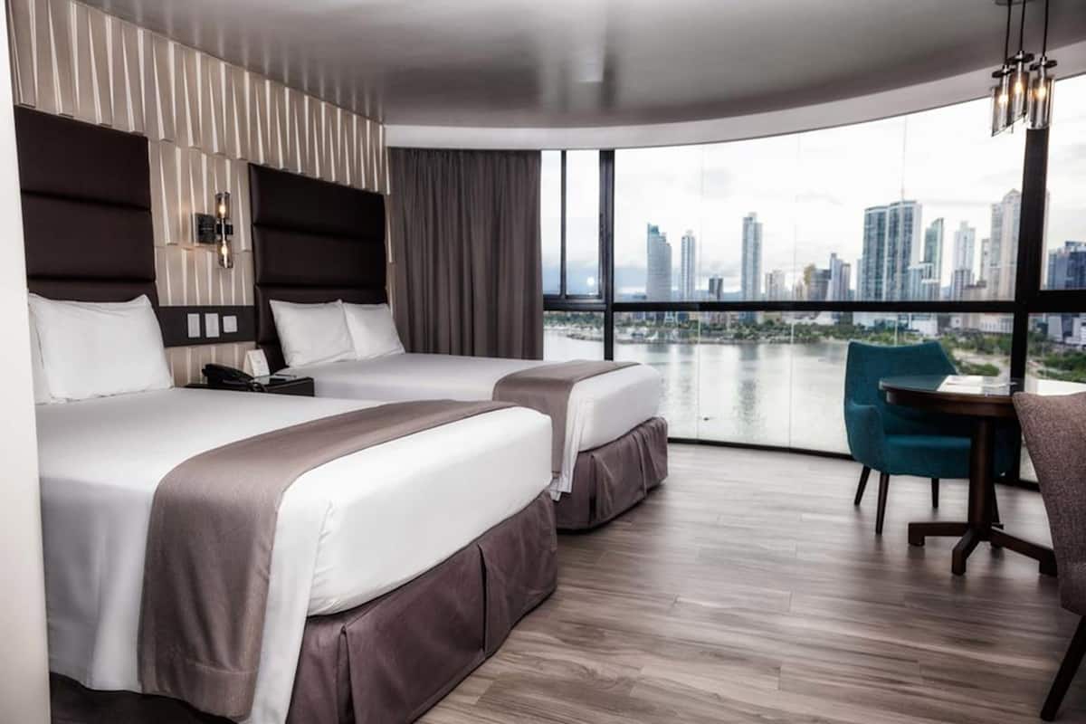 A hotel room with two beds, a desk, and a large window offering a city view.