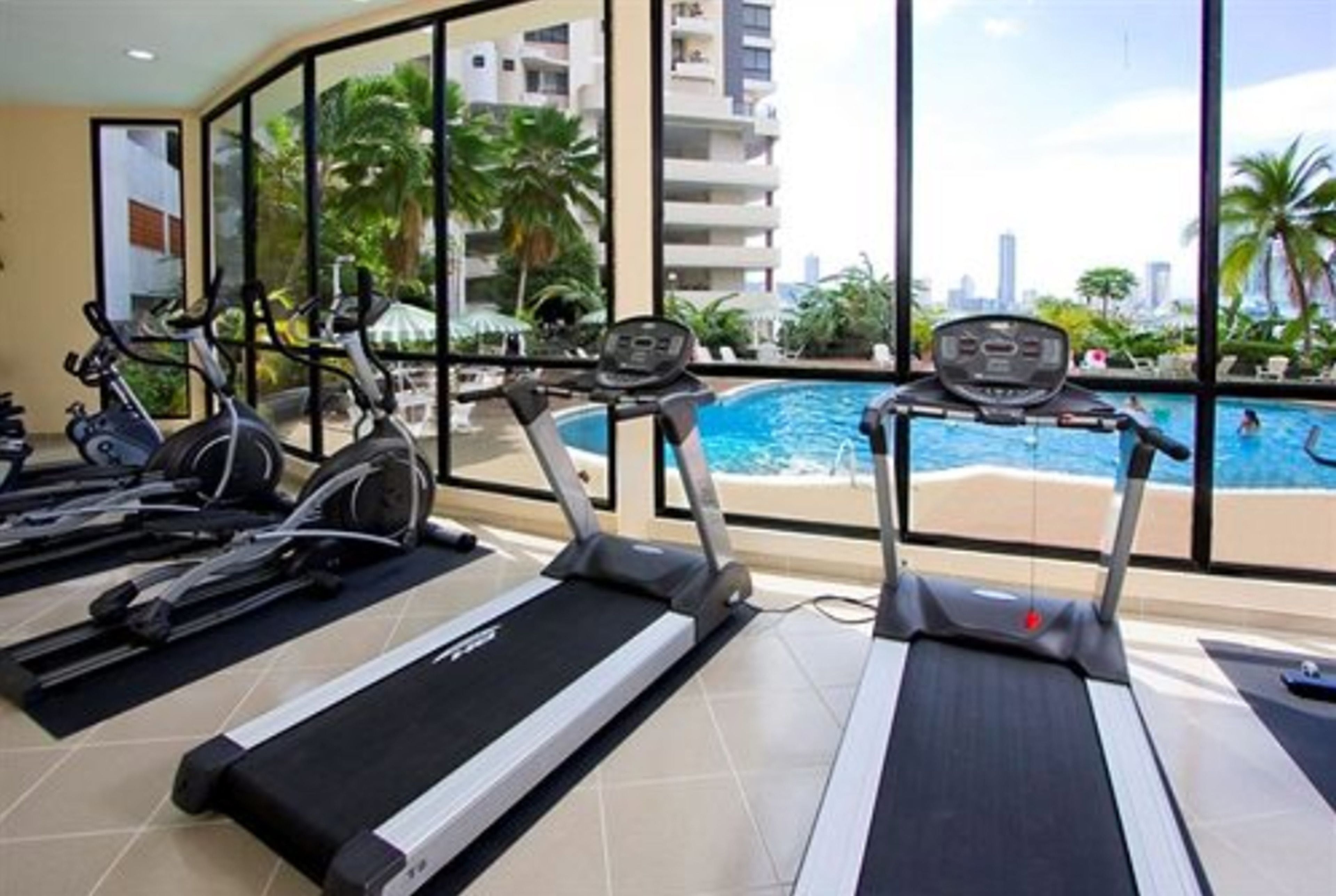 A gym with treadmills and stationary bikes, overlooking a pool and palm trees.
