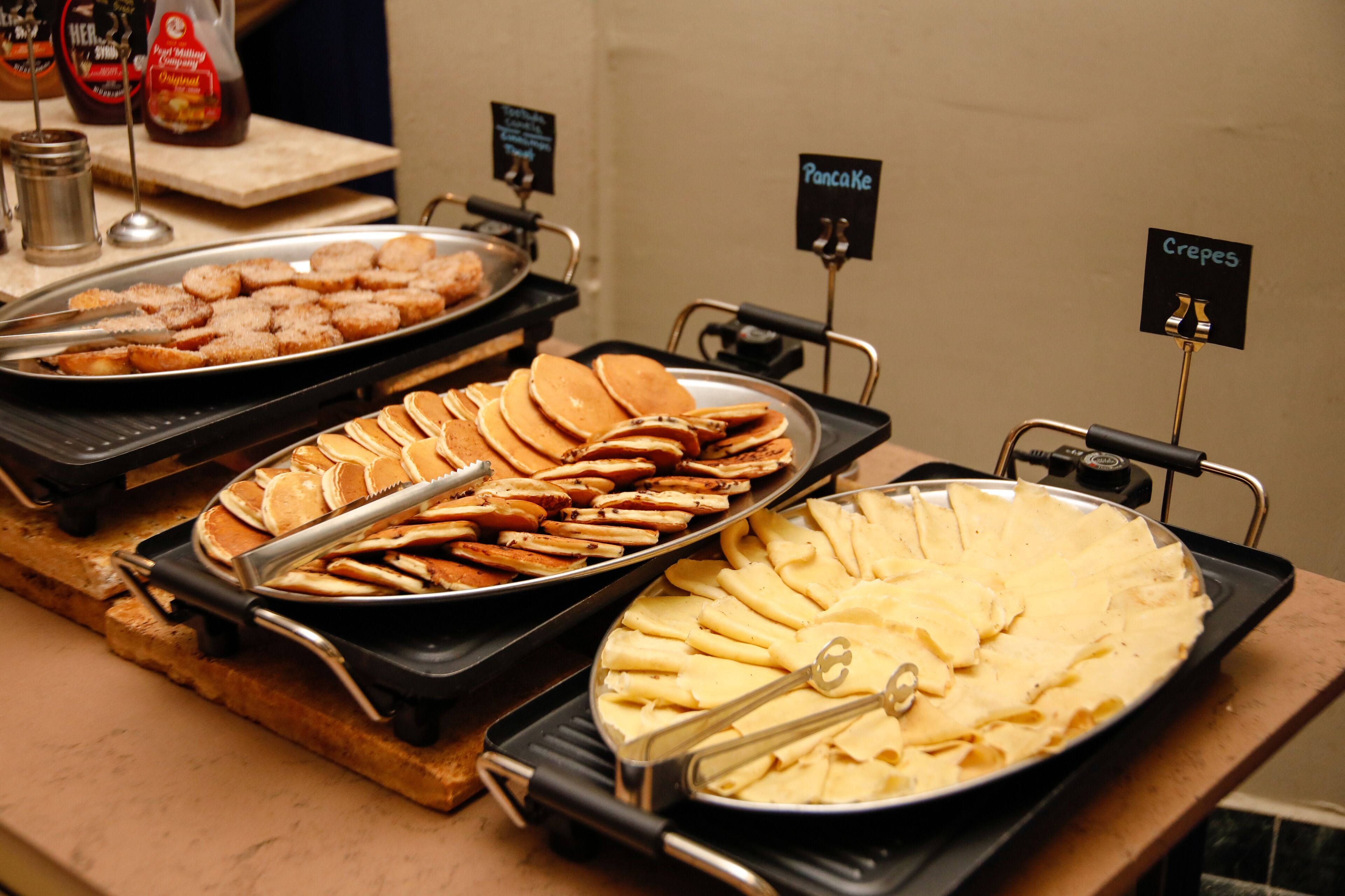A buffet with various food items like pancakes, waffles, and crepes, each labeled with their names.