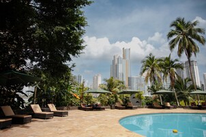Zona de piscina con sillones reclinables y sombrillas, con vistas despejadas a altos edificios al fondo y un ambiente tropical con palmeras.