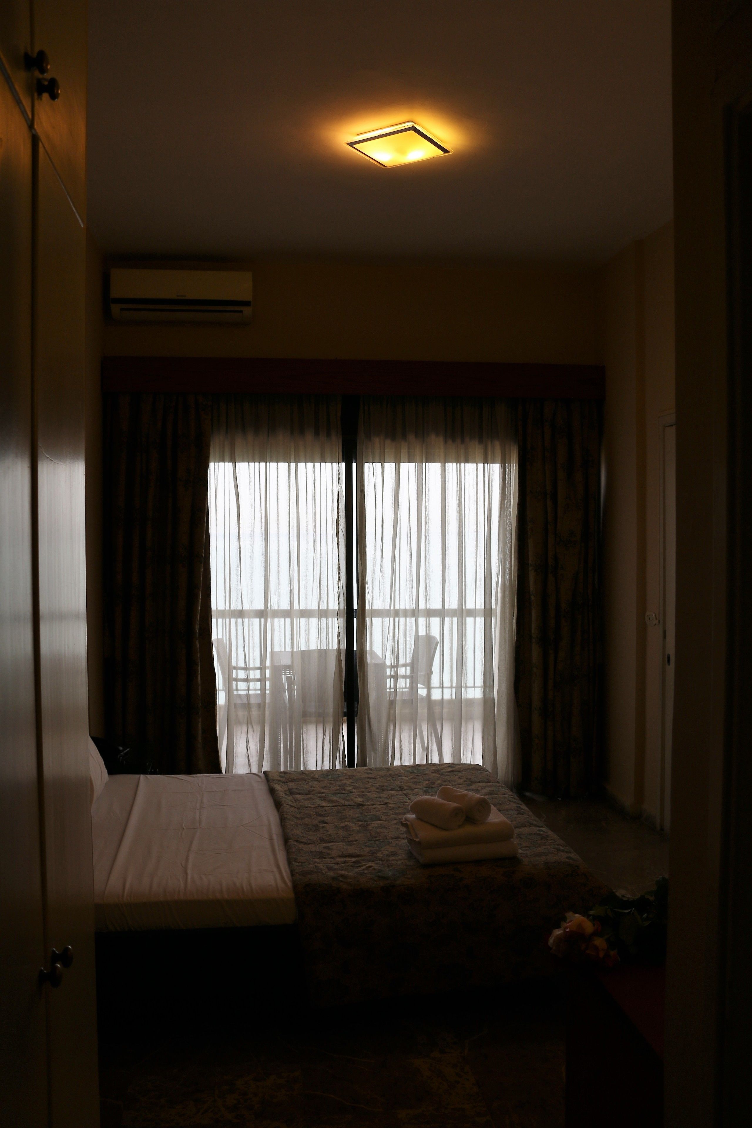 A dimly lit room with a bed, a small table with towels, and a chair near a window with sheer curtains.