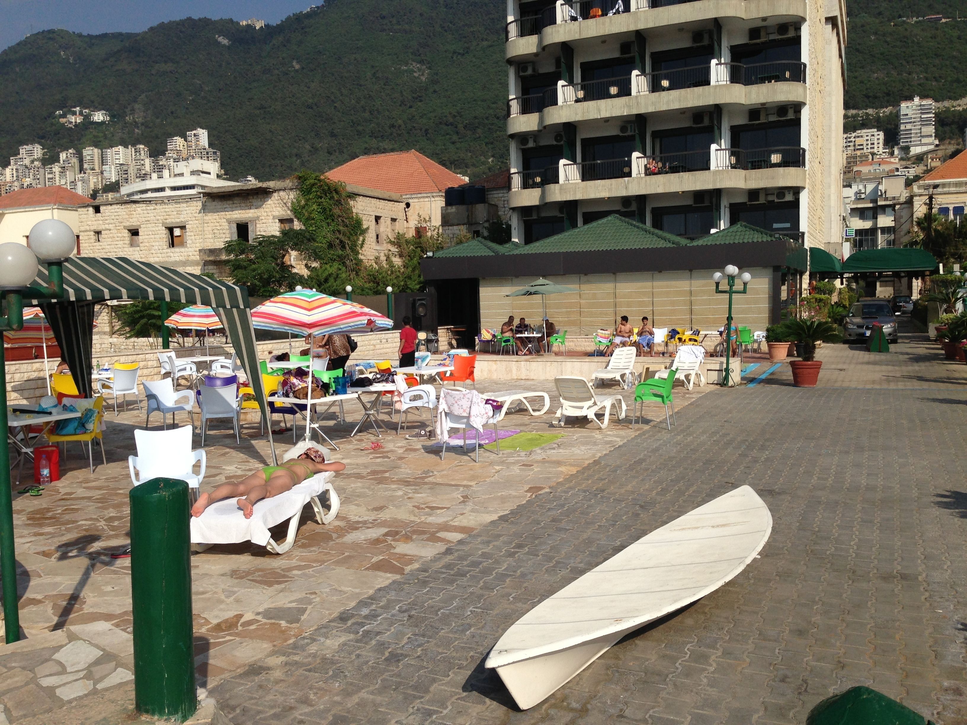 Un espacio al aire libre con mesas y sillas, una tabla de surf y un edificio con balcones al fondo.