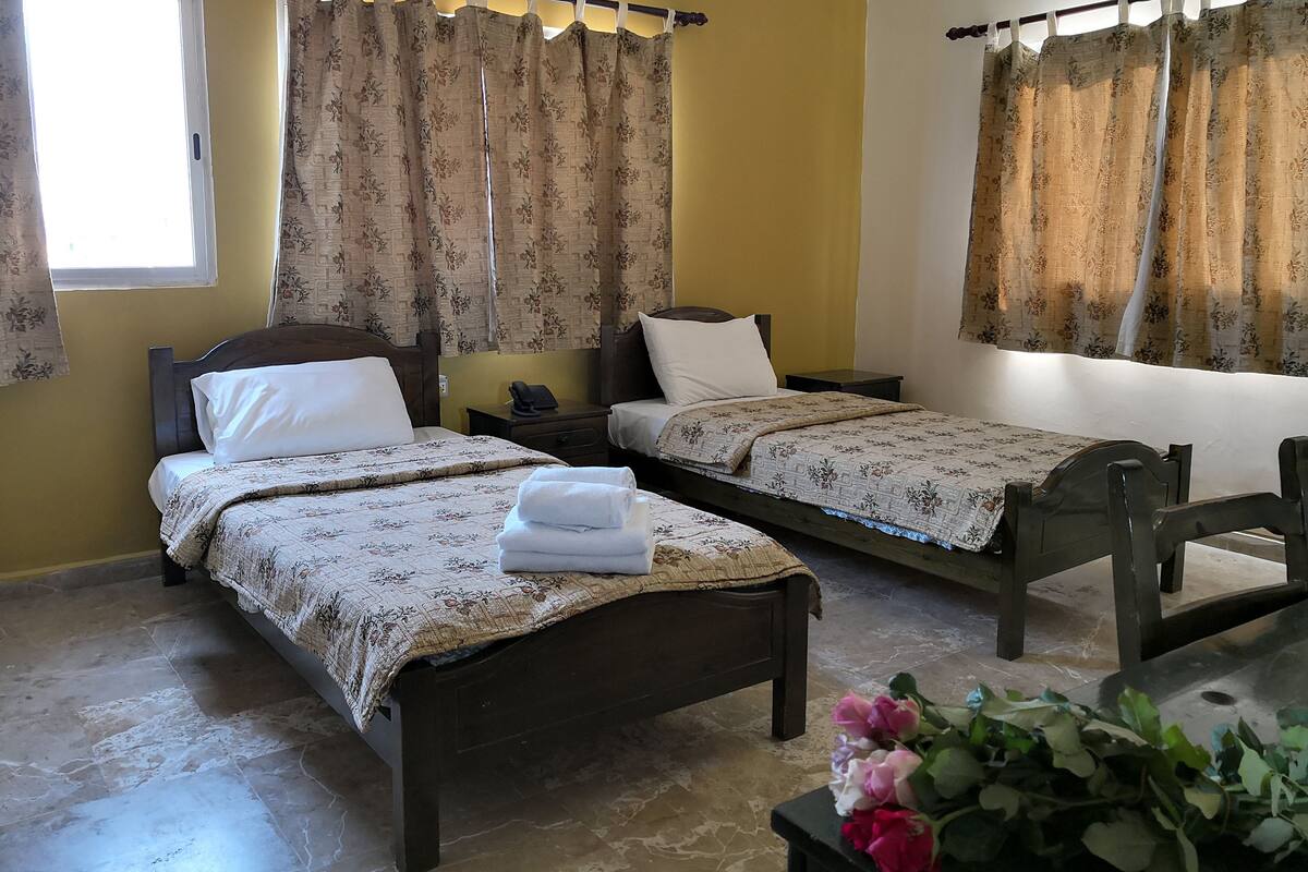 A hotel room with two single beds, a wooden headboard, a bedside table with a phone, and a small table with a bouquet of flowers.