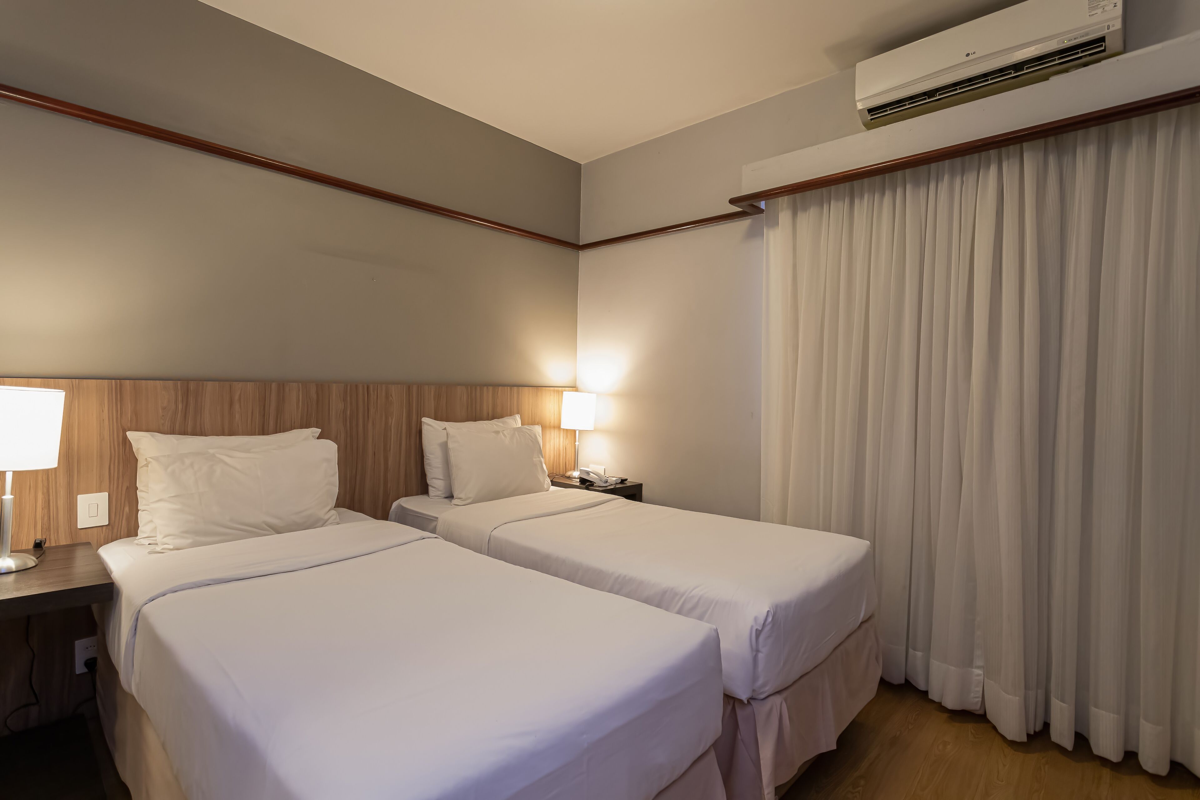 Ein Hotelzimmer mit zwei Betten, einem Nachttisch mit Lampe, einer Klimaanlage und einem großen Fenster mit Vorhängen.