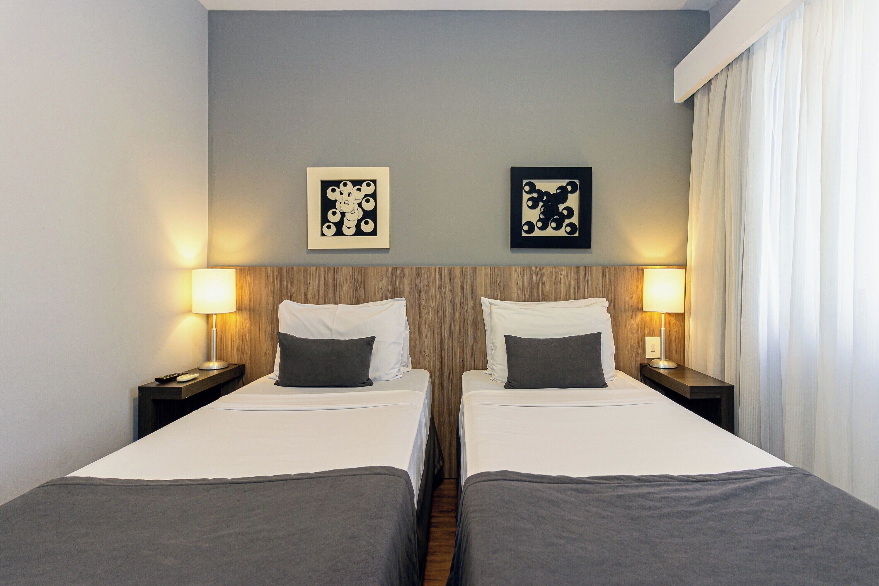 A hotel room with two beds, a wooden headboard, two bedside tables with lamps, and two framed pictures on the wall.
