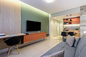 Un soggiorno moderno con una TV a schermo piatto, una parete decorativa in legno, una zona pranzo e una cucina sullo sfondo, e un divano grigio.