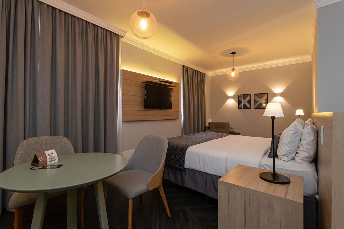 A hotel room with a bed, a desk, a chair, a TV, and two wall-mounted lamps.