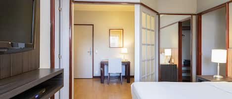 Quarto de hotel com cama, secretária, cadeira, televisão e abajur.